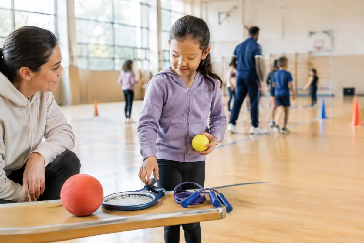 Un enfant découvre plusieurs équipements sportifs dans un gymnase, accompagné avec bienveillance par un parent.