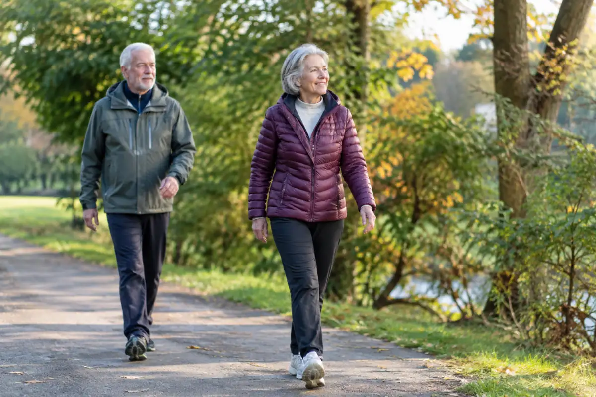 Deux adultes seniors marchent à allure modérée sur une allée plane de parc, avec des chaussures adaptées et une posture détendue.