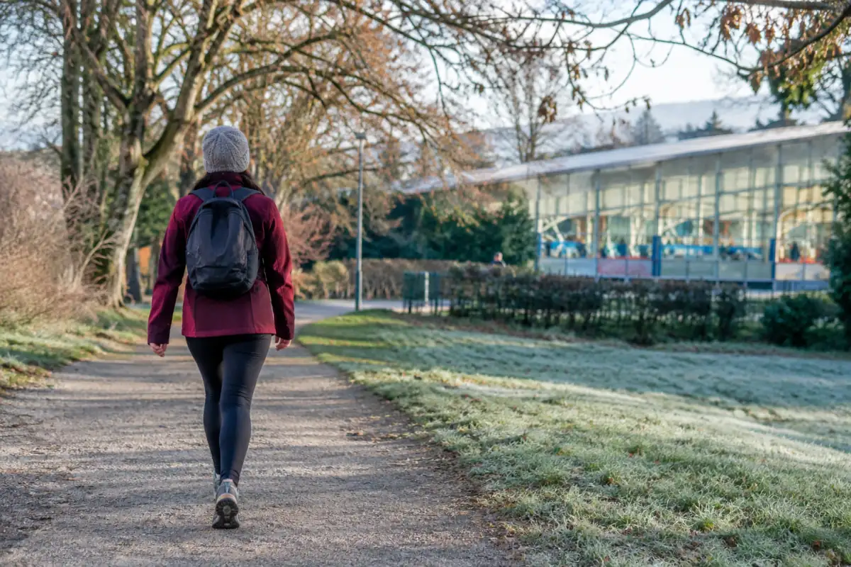 Personne marchant dans un parc en hiver, vêtue de couches adaptées, avec une salle de sport visible au loin.