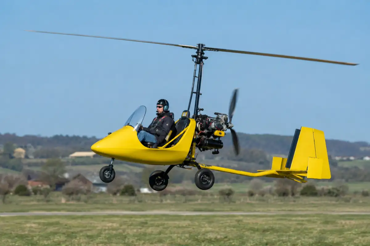 Un autogire moderne en vol au-dessus d’un aérodrome, avec son rotor libre et son hélice de propulsion visibles.