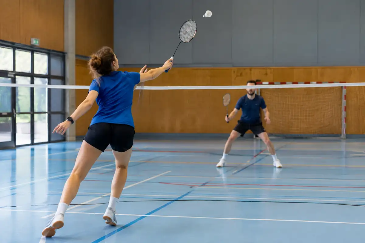 Échange de badminton en salle : une joueuse en fente frappe le volant au-dessus du filet sur un court réglementaire.