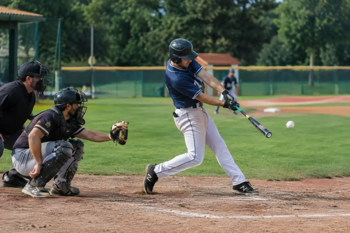 Un frappeur équipé prend son élan au marbre pendant qu’un receveur attend la balle sur un terrain de baseball.