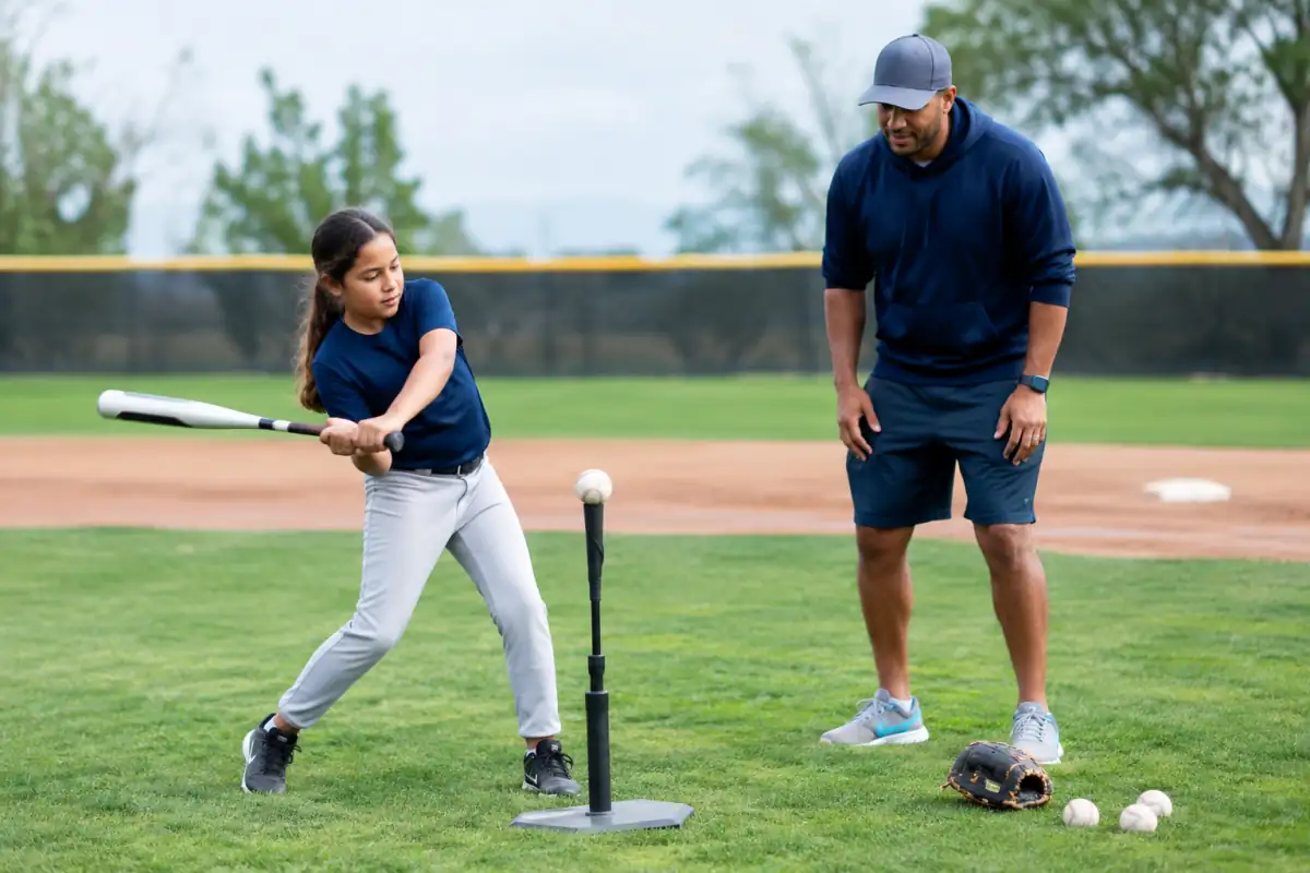 Une débutante travaille une frappe au tee sur un terrain de baseball, sous le regard attentif de son entraîneur.