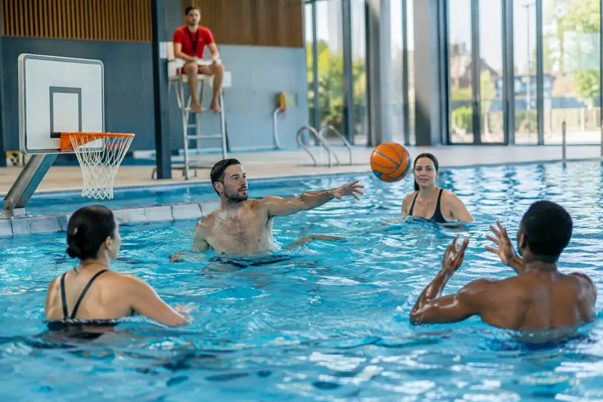 Des joueurs s’échangent un ballon lors d’une partie de basket-ball aquatique dans une piscine avec panier au bord du bassin.