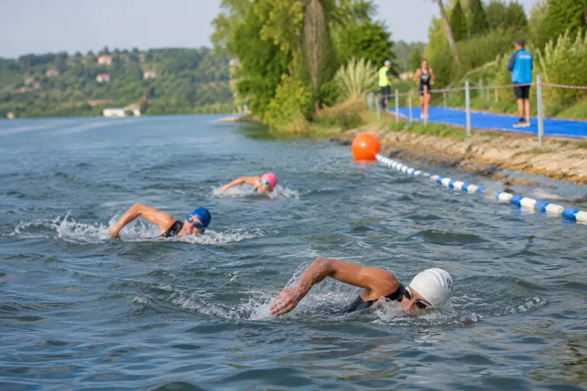 Des concurrents nagent en eau libre sur le segment natation d’une épreuve de biathle organisée près d’un parcours de course.