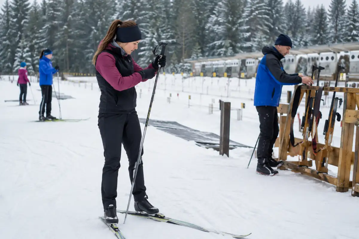 Débutant en skis de skating vérifiant ses bâtons près d’un pas de tir où la carabine reste rangée sous l’encadrement du club.