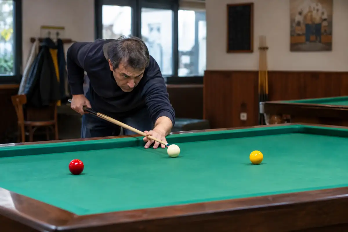 Joueur visant un carambolage sur une table sans poches avec trois billes pendant une séance de billard français en club.