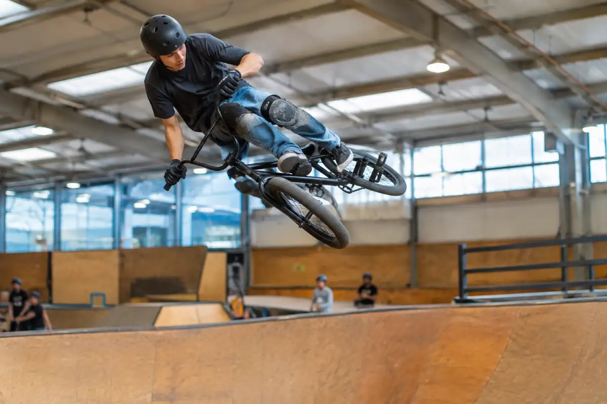 Un pilote en BMX park réalise un saut incliné au-dessus d’une rampe, avec casque et protections dans un skatepark couvert.