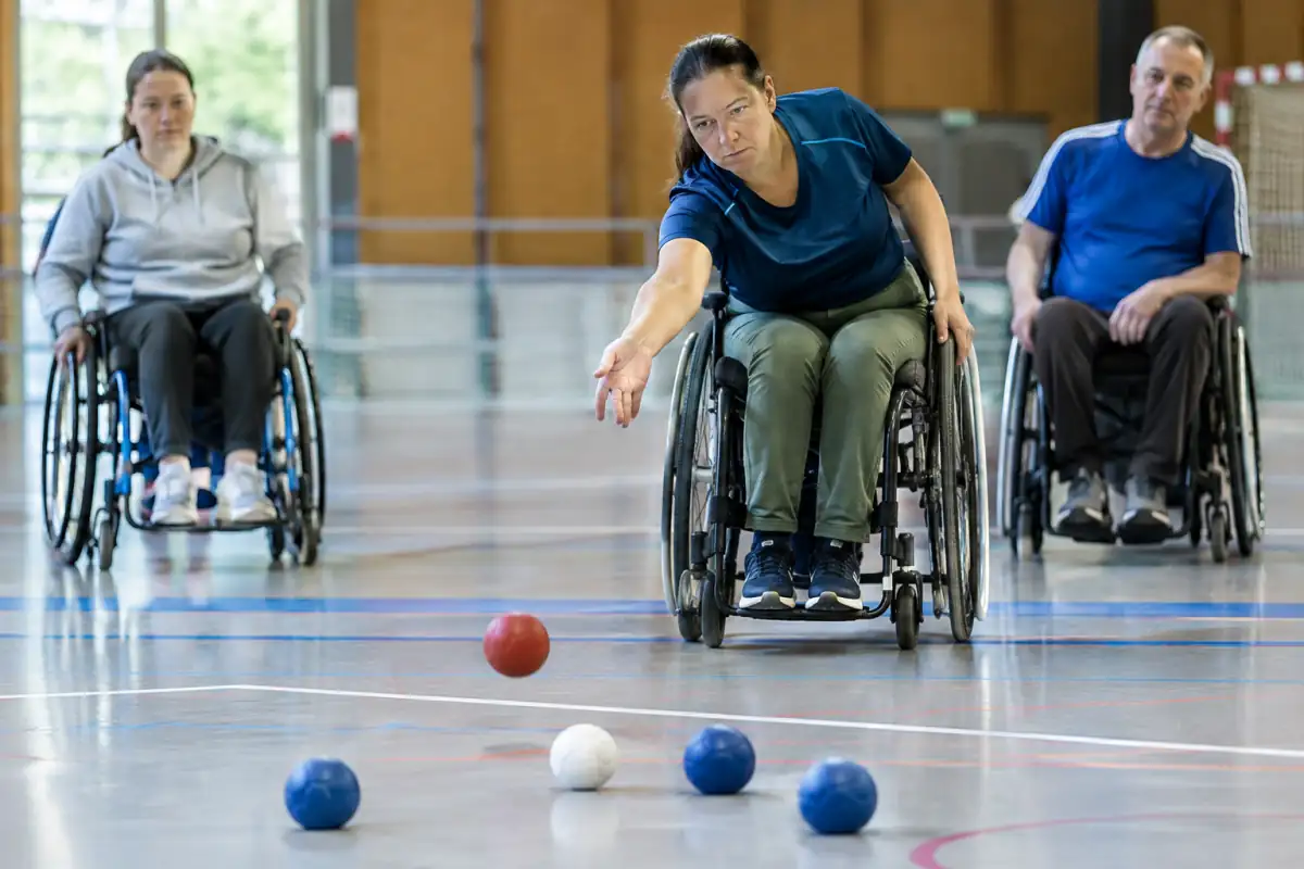 Une joueuse de boccia en fauteuil lance une balle rouge vers le jack blanc sur un terrain intérieur, tandis que son adversaire observe.