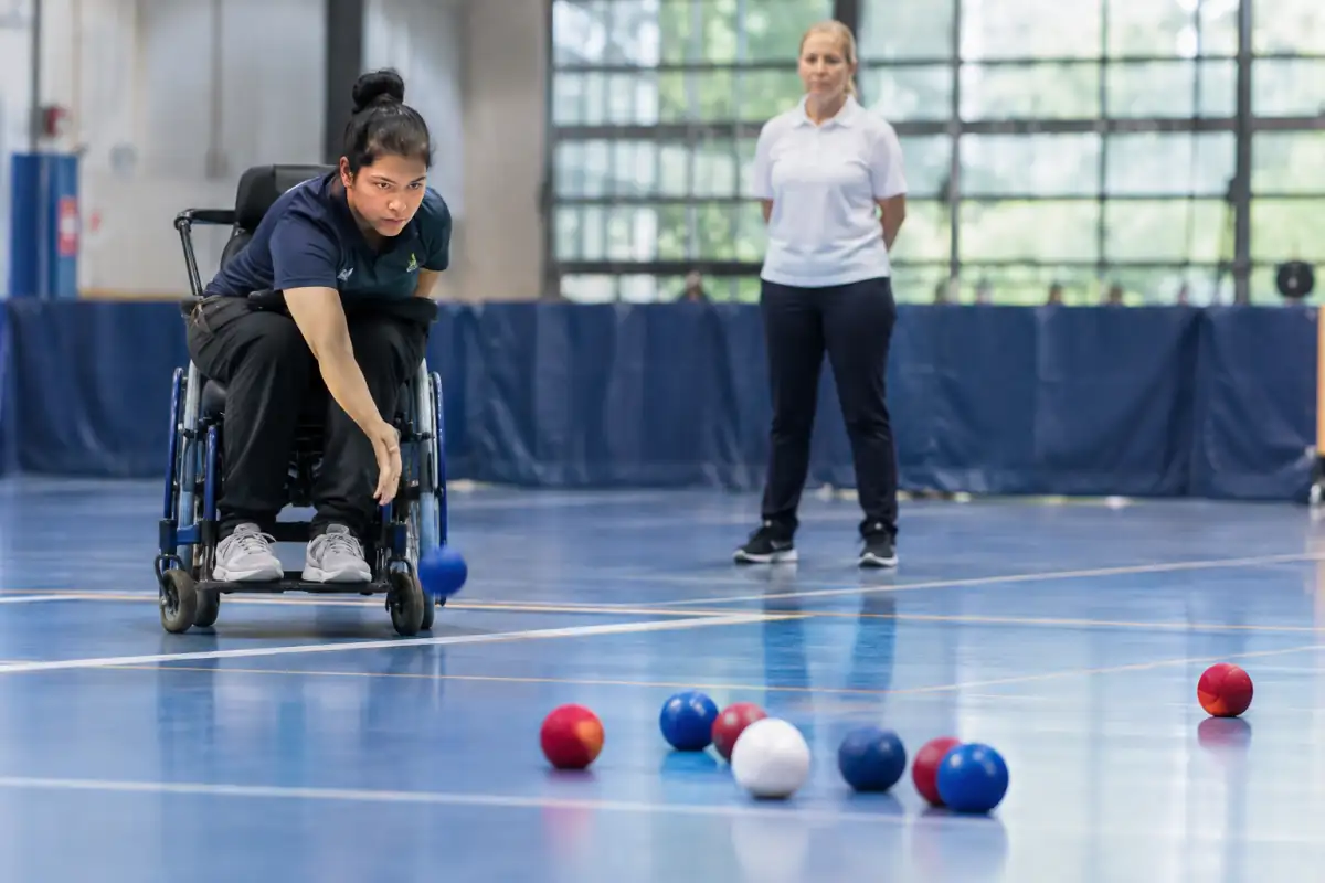 Joueur de boccia prêt à lancer une balle rouge vers le jack blanc, au milieu de balles bleues sur un terrain intérieur balisé.