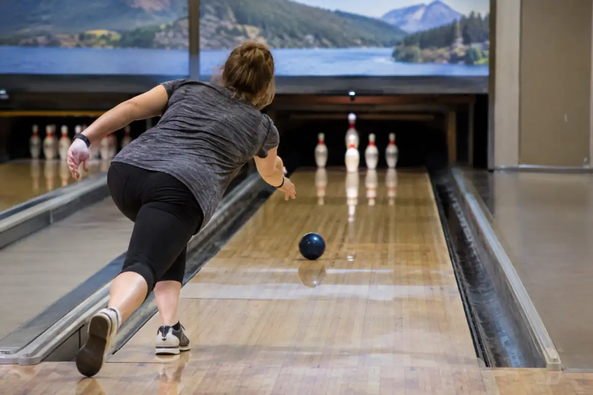 Un joueur lance une petite boule sans trous sur une piste de bowling à cinq quilles, disposées en V au fond.