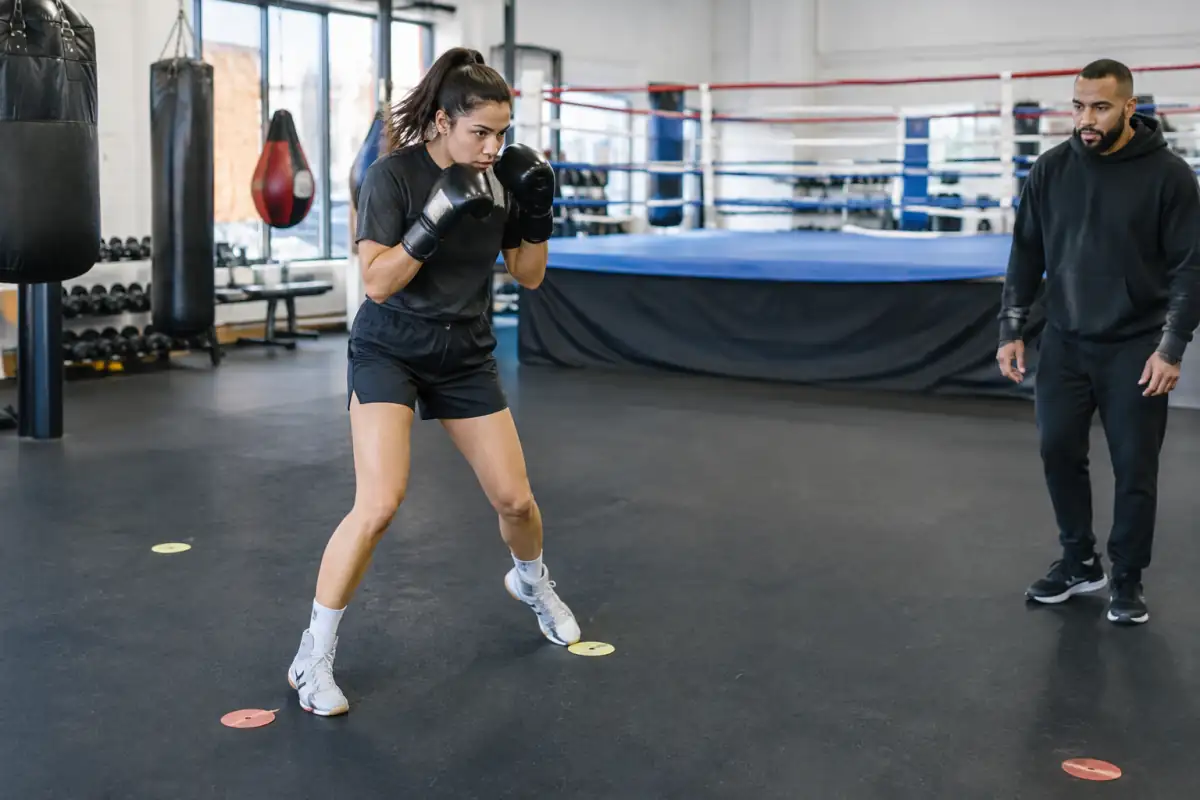 Débutant en boxe anglaise travaillant ses déplacements en garde autour de repères au sol, sous l’observation d’un entraîneur.