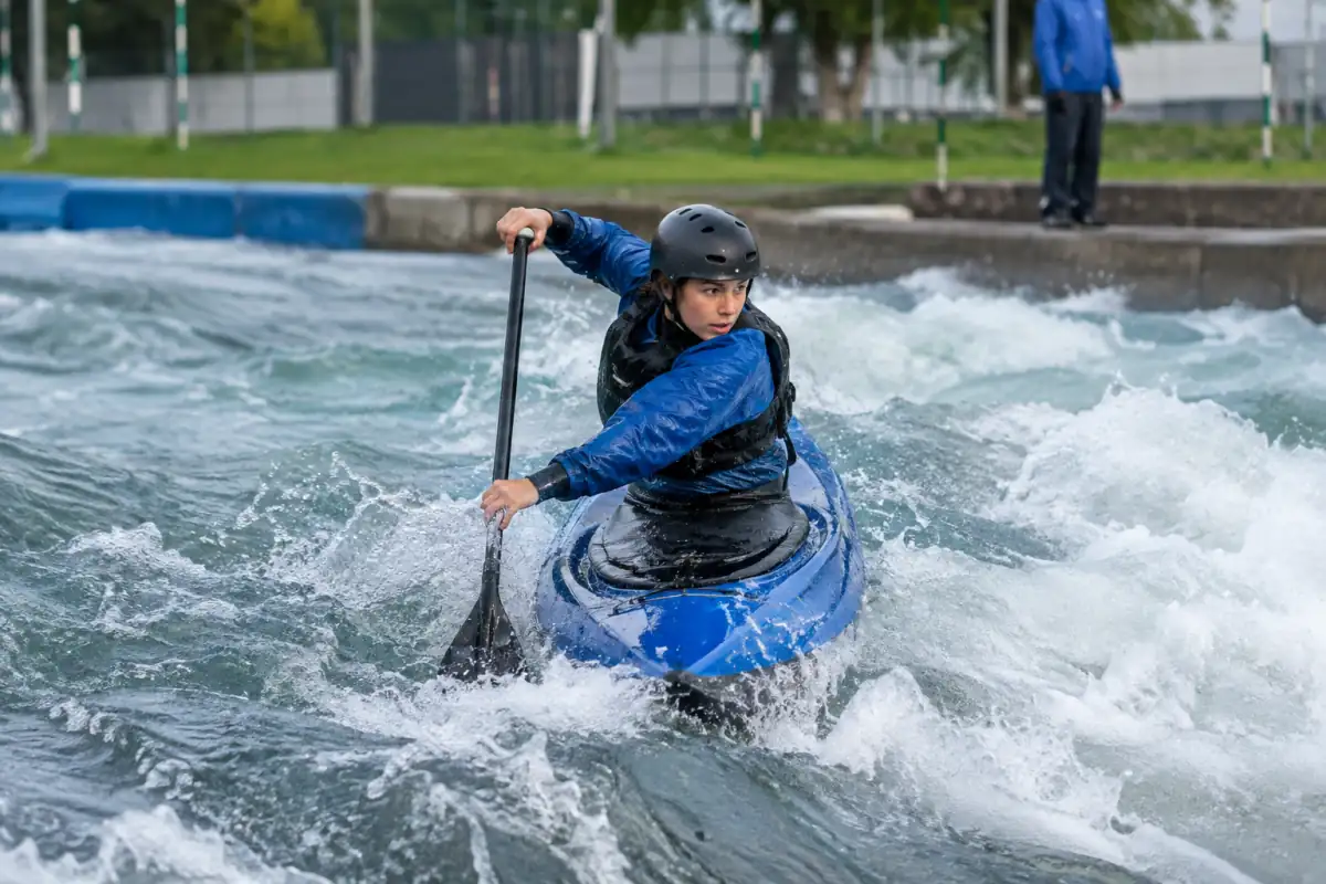 Un pagayeur à genoux réalise une figure en canoë freestyle sur une vague d’eau vive, avec une pagaie simple.