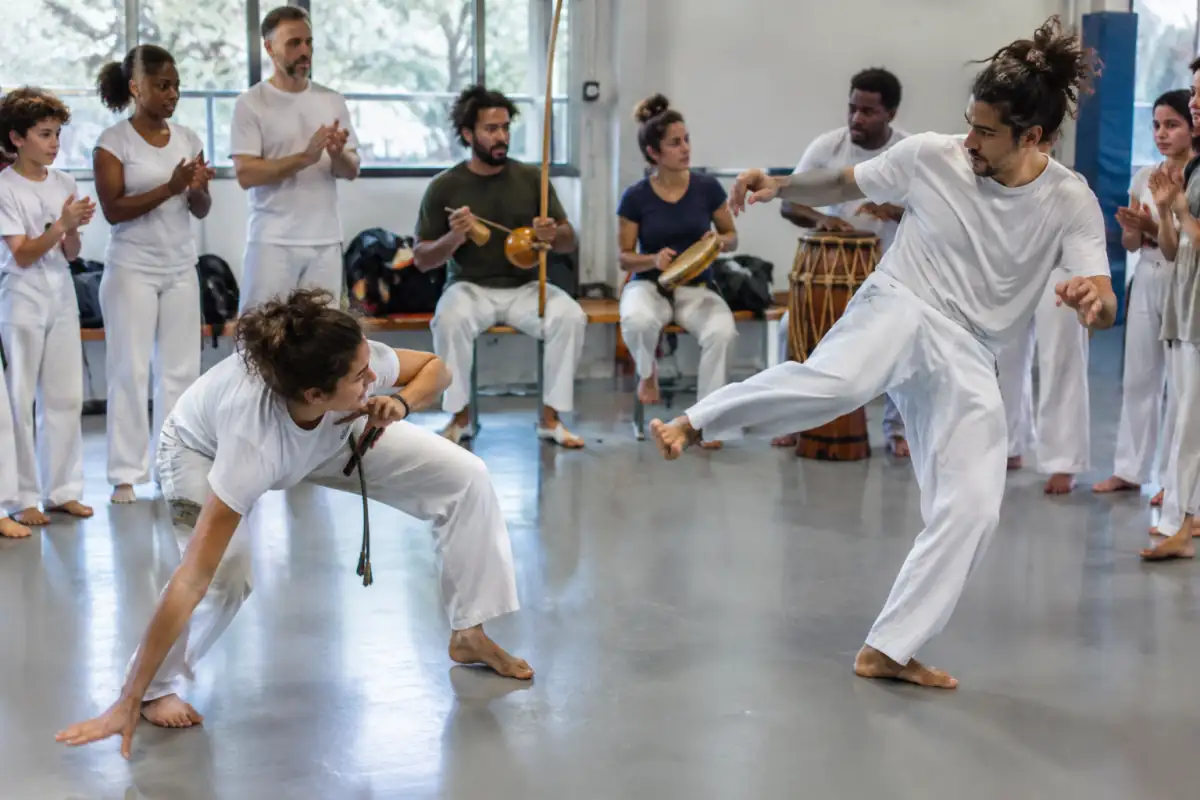 Deux capoeiristes jouent dans une roda, entre esquives et ginga, tandis que les musiciens donnent le rythme au berimbau.