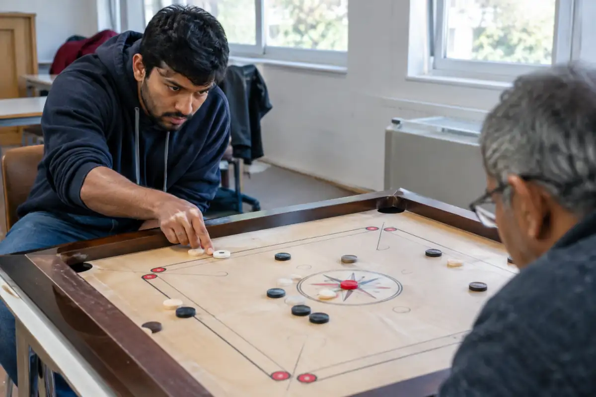Une joueuse propulse du doigt le percuteur sur un plateau de carrom, face aux palets et aux poches d’un club associatif.