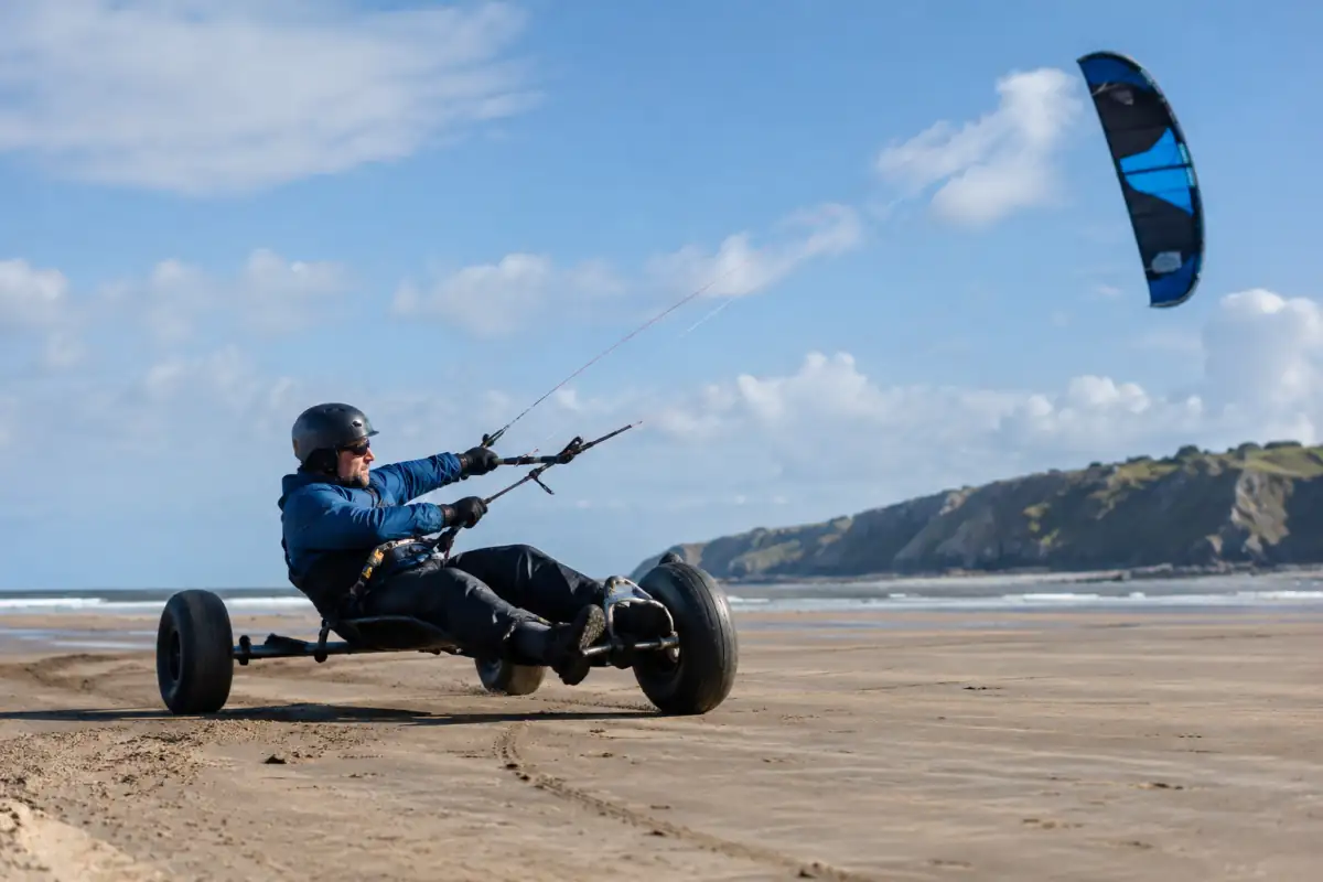 Un pratiquant pilote une aile de traction depuis un buggy à trois roues sur une plage dégagée, casque et harnais.