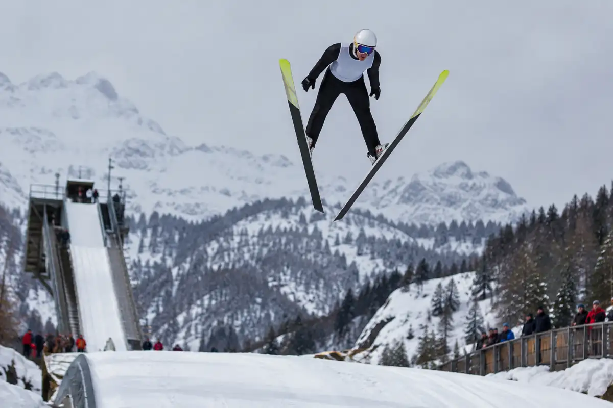 Athlète de combiné nordique en plein saut, skis en V au-dessus d’un tremplin enneigé dans les Alpes françaises.