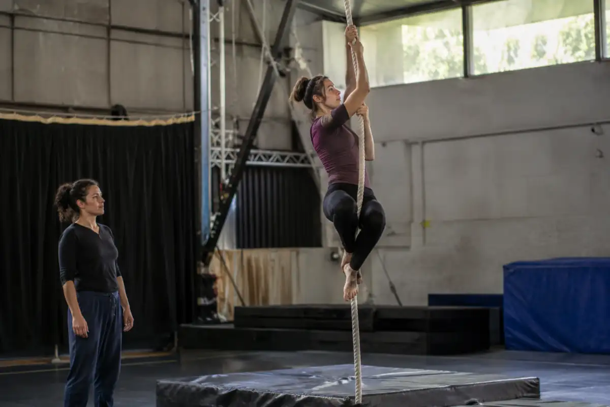 Une élève de cirque travaille une montée contrôlée sur corde aérienne au-dessus de tapis, sous le regard d’un professeur.