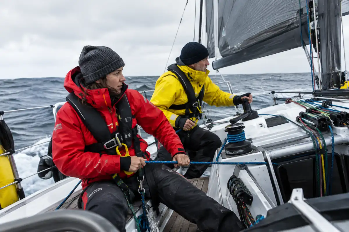 Deux équipiers en tenue hauturière règlent les voiles d’un monocoque engagé dans une course au large.