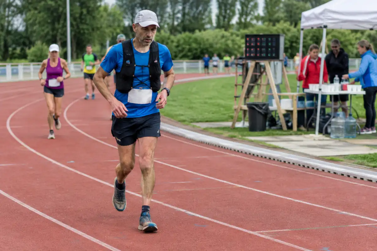 Coureur en tenue technique qui avance sur une piste lors d’une course de 6 jours, avec d’autres participants et un ravitaillement en arrière-plan.