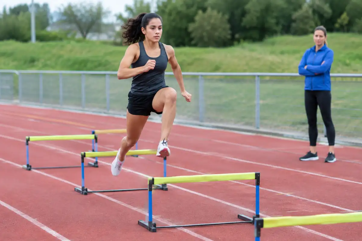 Débutant en athlétisme travaillant le franchissement de haies basses sur une piste, sous la supervision d’un entraîneur.