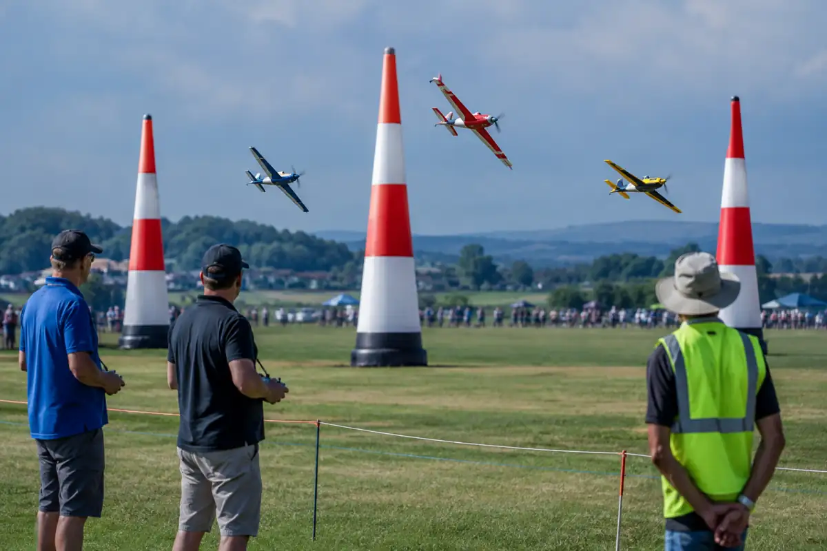 Trois avions modèles de course contournent des pylônes sur un terrain d’aéromodélisme, devant les pilotes.