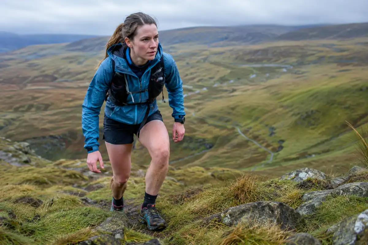 Coureuse de fell running en veste imperméable et chaussures à crampons sur une pente herbeuse de lande britannique.
