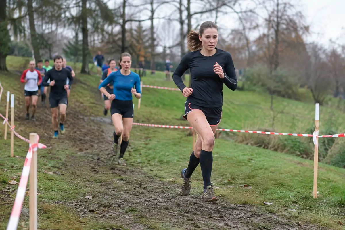 Coureuse de cross-country relançant sur une portion boueuse d’un parcours en boucle, avec d’autres participants dans un parc d’automne.