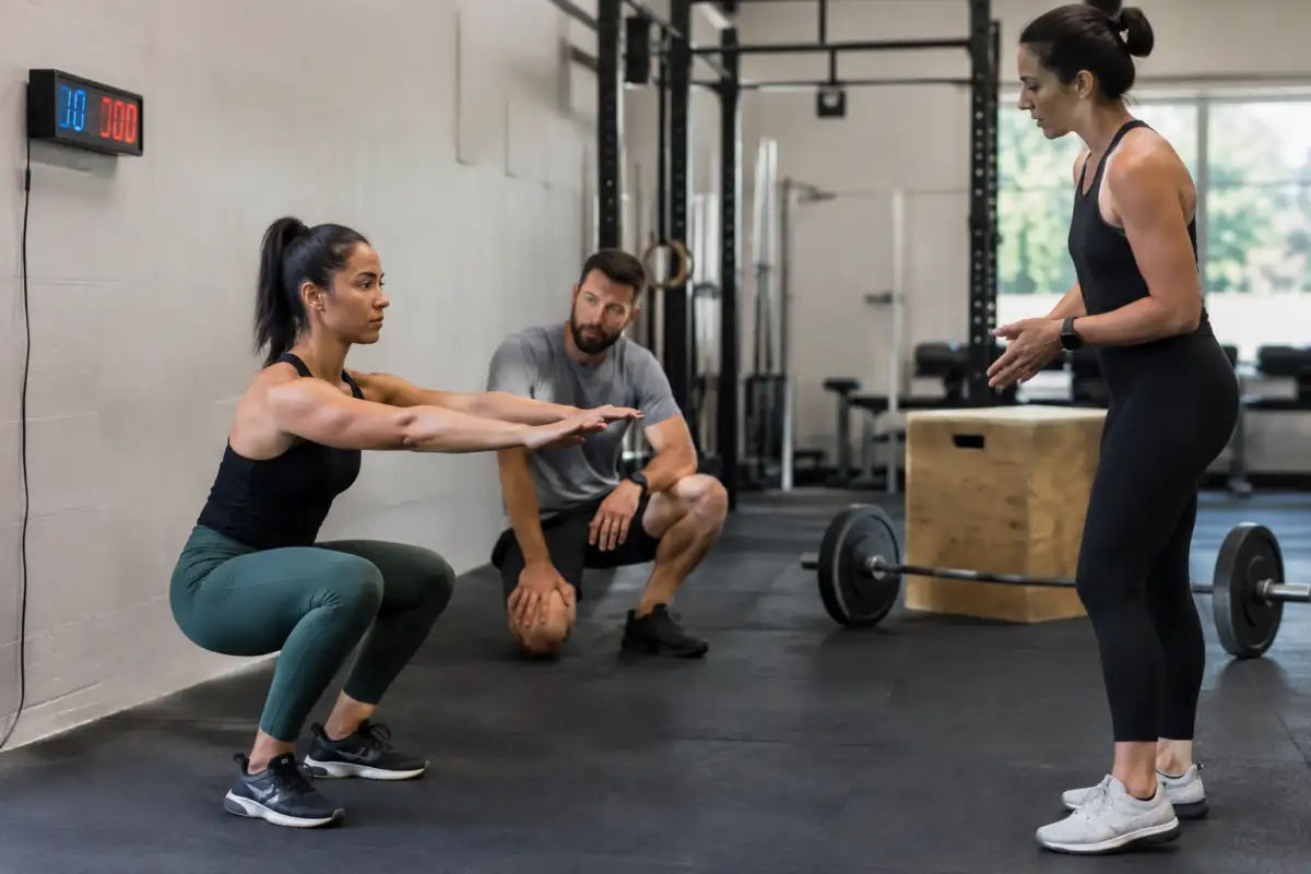 Un coach montre une position de squat dans une salle de cross-training pendant qu’un minuteur rythme un EMOM.