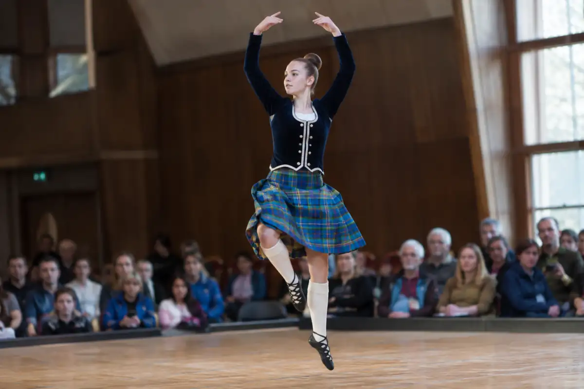 Danseuse exécutant un saut de Highland dance en kilt tartan et ghillies sur une scène lors d’une compétition.