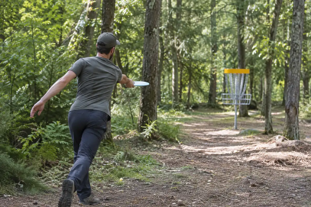 Un joueur lance un disque sur un parcours boisé de disc golf, avec un panier métallique visible entre les arbres.
