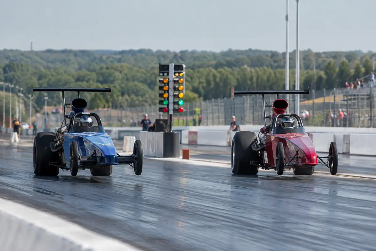 Deux dragsters s’élancent côte à côte au départ d’une piste aménagée, pilotes casqués et feux de signalisation visibles.