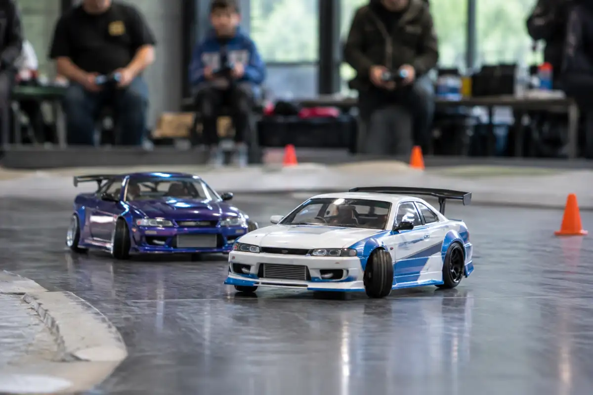 Deux voitures de drift radiocommandé en tandem négocient un virage sur une piste lisse balisée par des cônes.