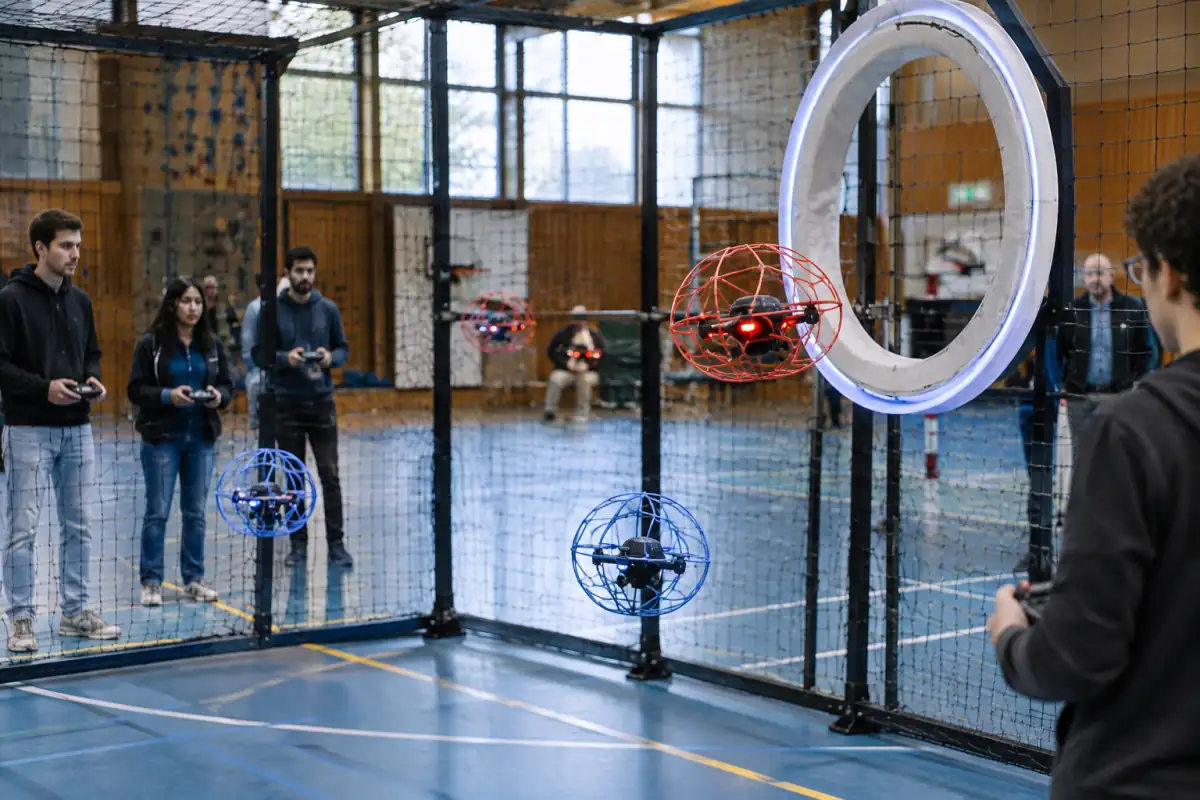 Match de drone soccer en salle : un drone protégé approche de l’anneau de but pendant que les pilotes dirigent leurs appareils.