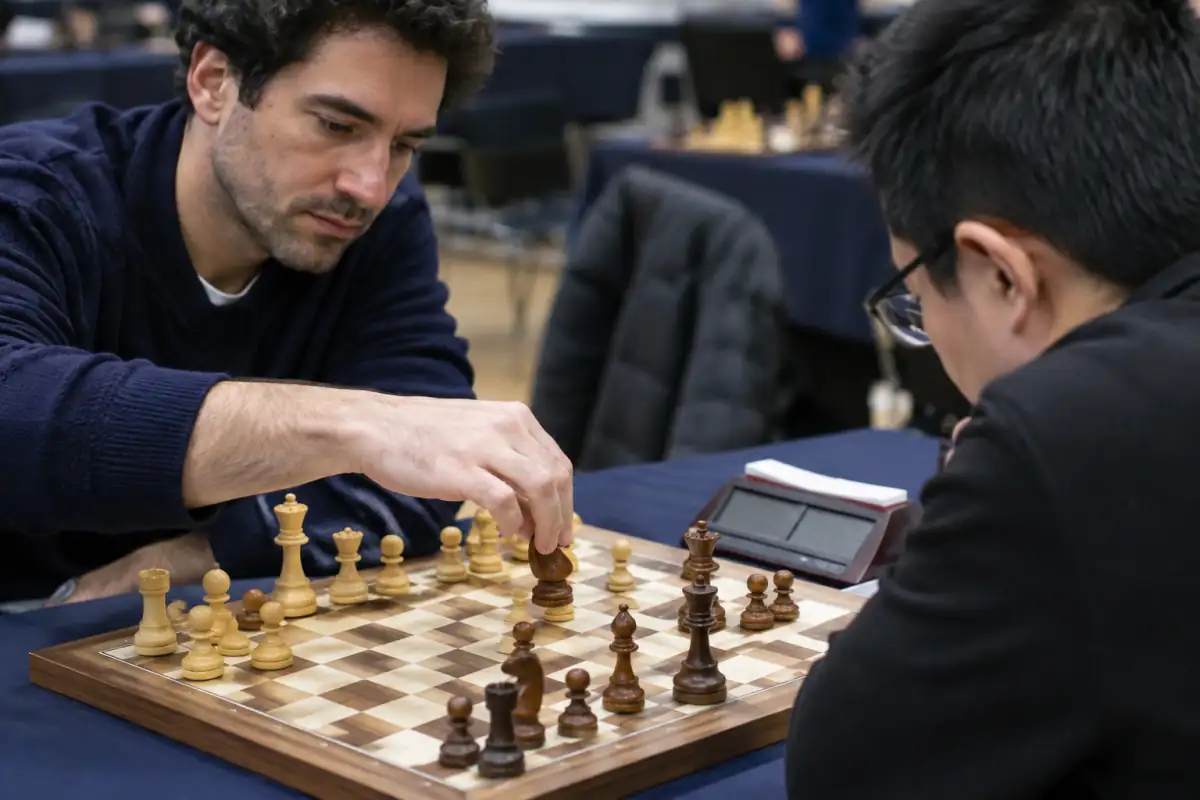 Deux joueurs concentrés disputent un blitz sur un échiquier, une main prête à déplacer un cavalier près d’une pendule numérique.