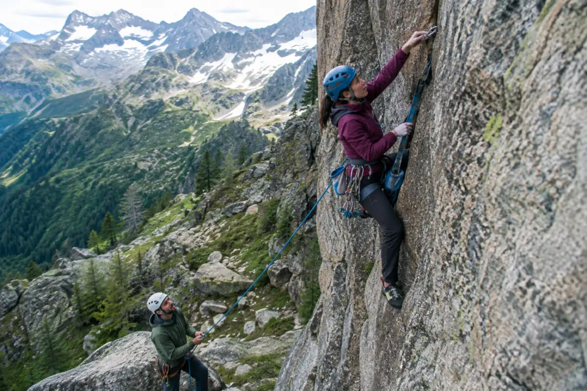 Un grimpeur en escalade artificielle monte dans un étrier sur une paroi rocheuse, assuré par un partenaire à la corde.