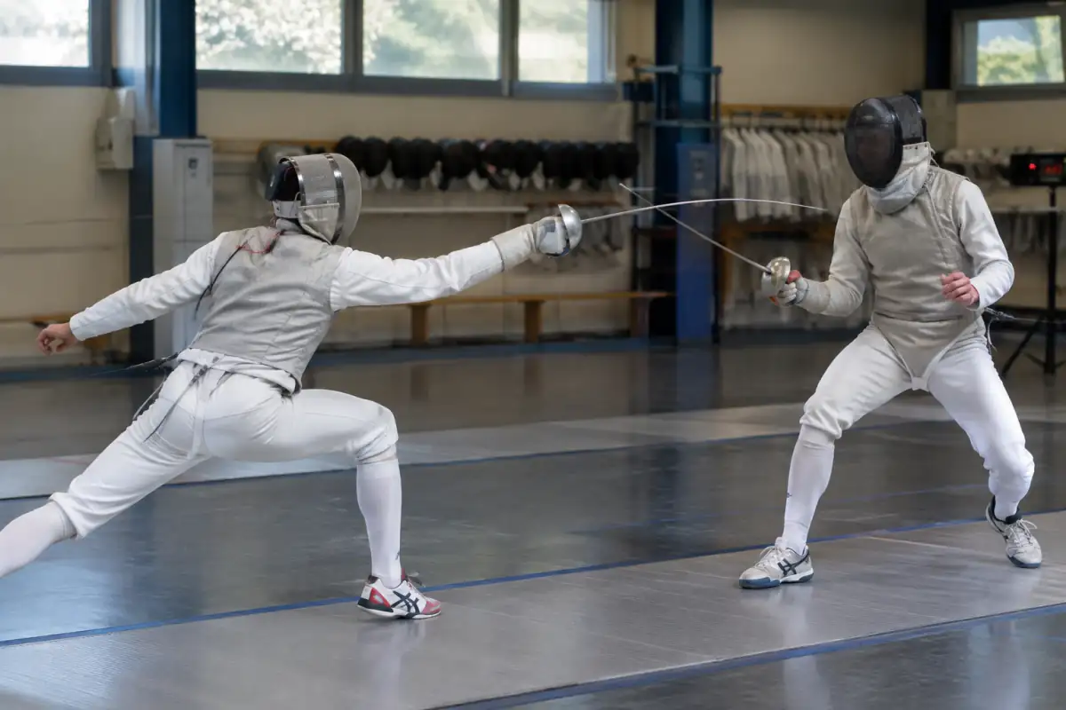 Deux sabreurs en tenue conductrice s’affrontent sur une piste lors d’un entraînement d’escrime au sabre en salle.