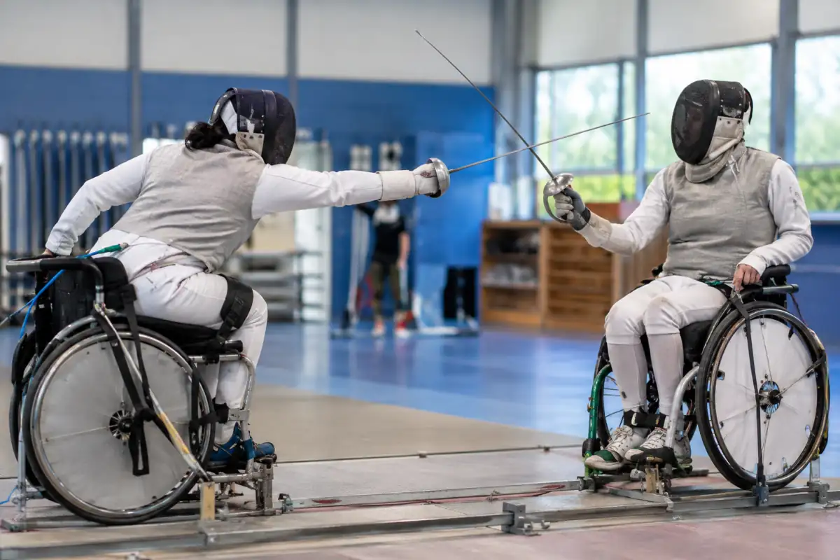 Deux tireurs en escrime fauteuil s’affrontent à l’épée, leurs fauteuils fixés au cadre dans une salle de club.