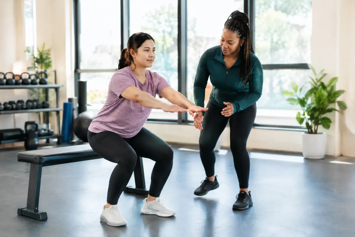 Une débutante effectue un squat vers un banc stable en salle, tandis qu’un coach observe sa posture et la guide.