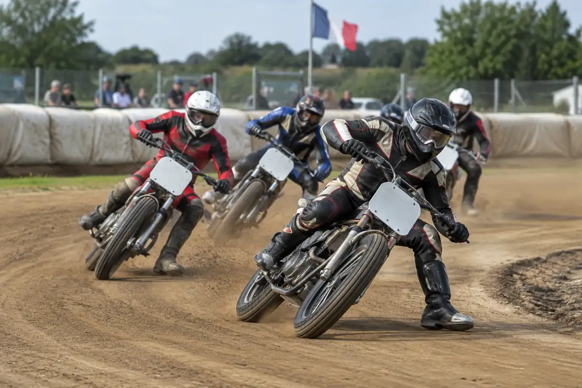 Pilotes de flat track moto en glisse dans un virage à gauche sur une piste ovale en terre.