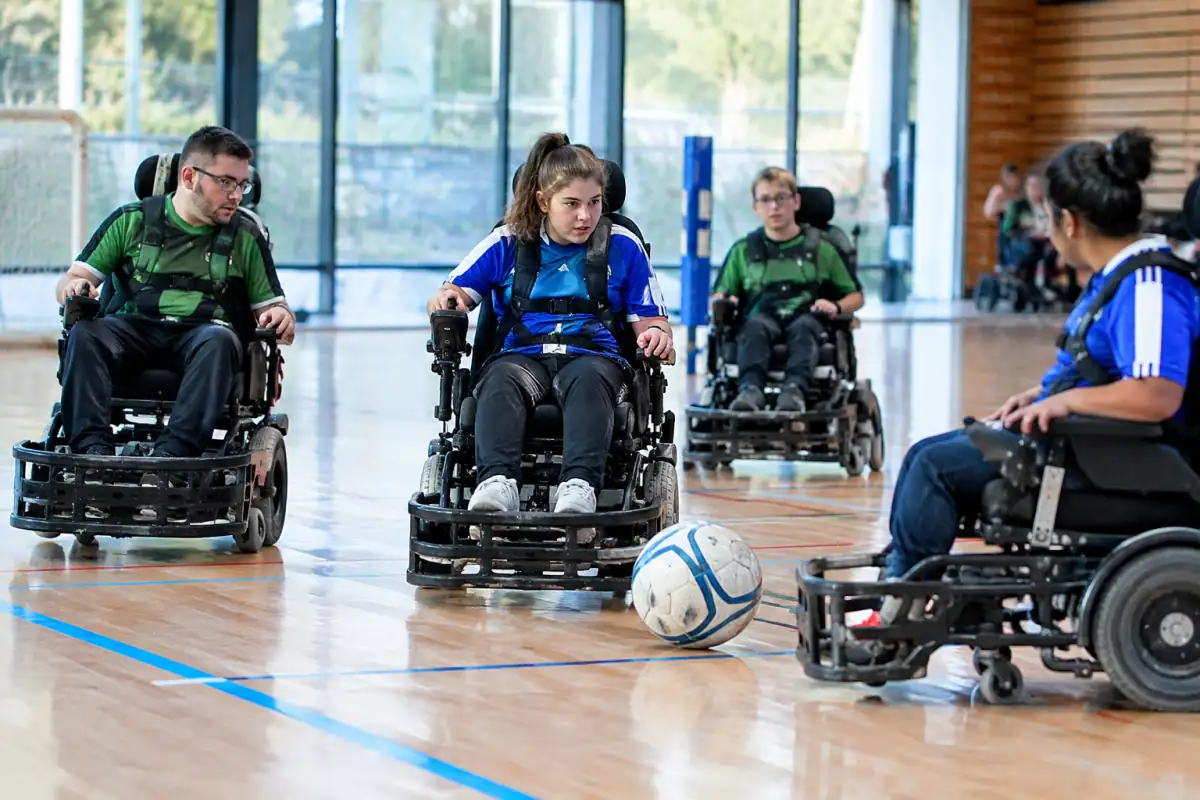 Joueurs de foot fauteuil électrique disputent un ballon surdimensionné dans un gymnase avec leurs pare-chocs avant.