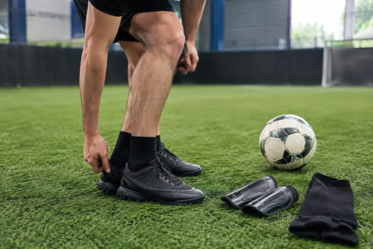 Joueur vérifiant des chaussures TF à petits crampons sur gazon synthétique, avec protège-tibias et chaussettes montantes.