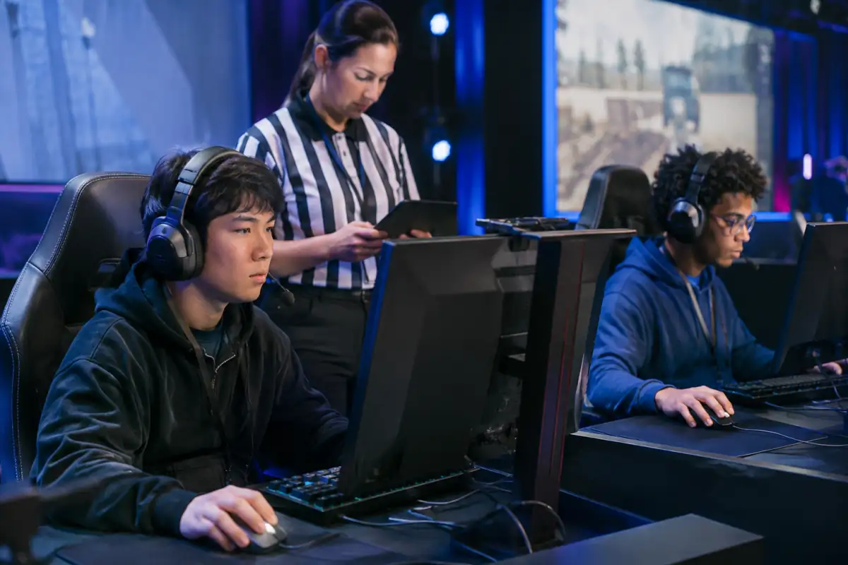 Un arbitre vérifie une tablette près de joueurs concentrés devant leur écran lors d’un match de FPS en esport.