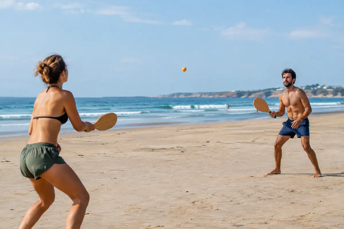 Deux partenaires échangent une balle avec des palettes en bois lors d’une partie de frescobol sur une plage dégagée.