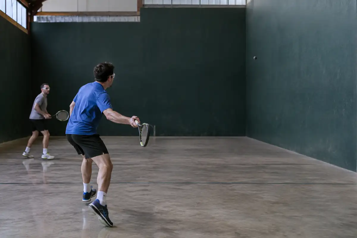 Échange de frontenis dans un fronton couvert, avec raquettes de tennis, balle en caoutchouc et lunettes de protection.