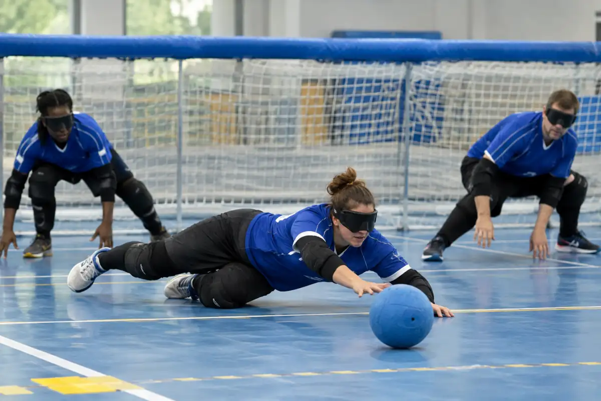 Des joueurs de goalball plongent au sol pour bloquer un ballon sonore sur un terrain intérieur aux lignes tactiles.