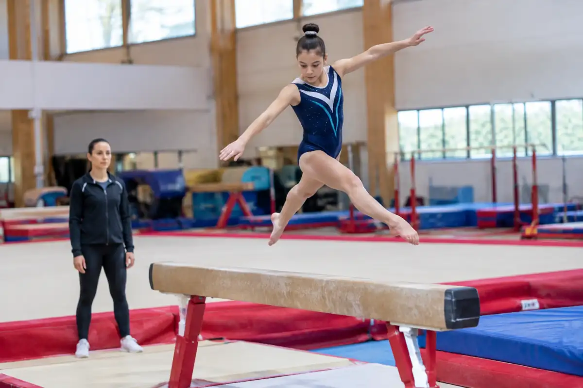 Gymnastique artistique féminine : une gymnaste réalise un élément acrobatique sur une poutre dans une salle équipée de tapis.