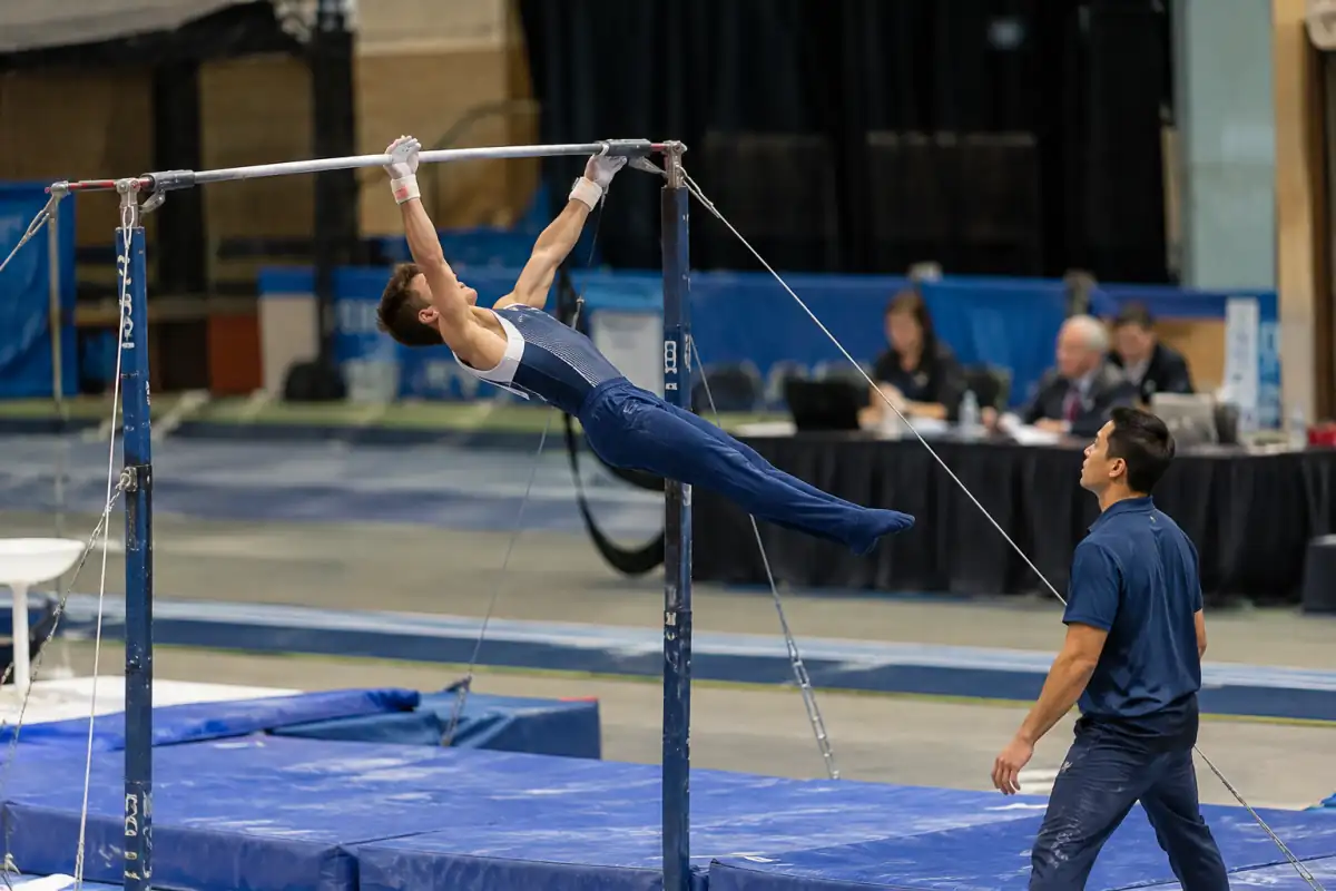 Gymnaste masculin en plein lâcher à la barre fixe, sous le regard attentif d’un pareur et des juges.