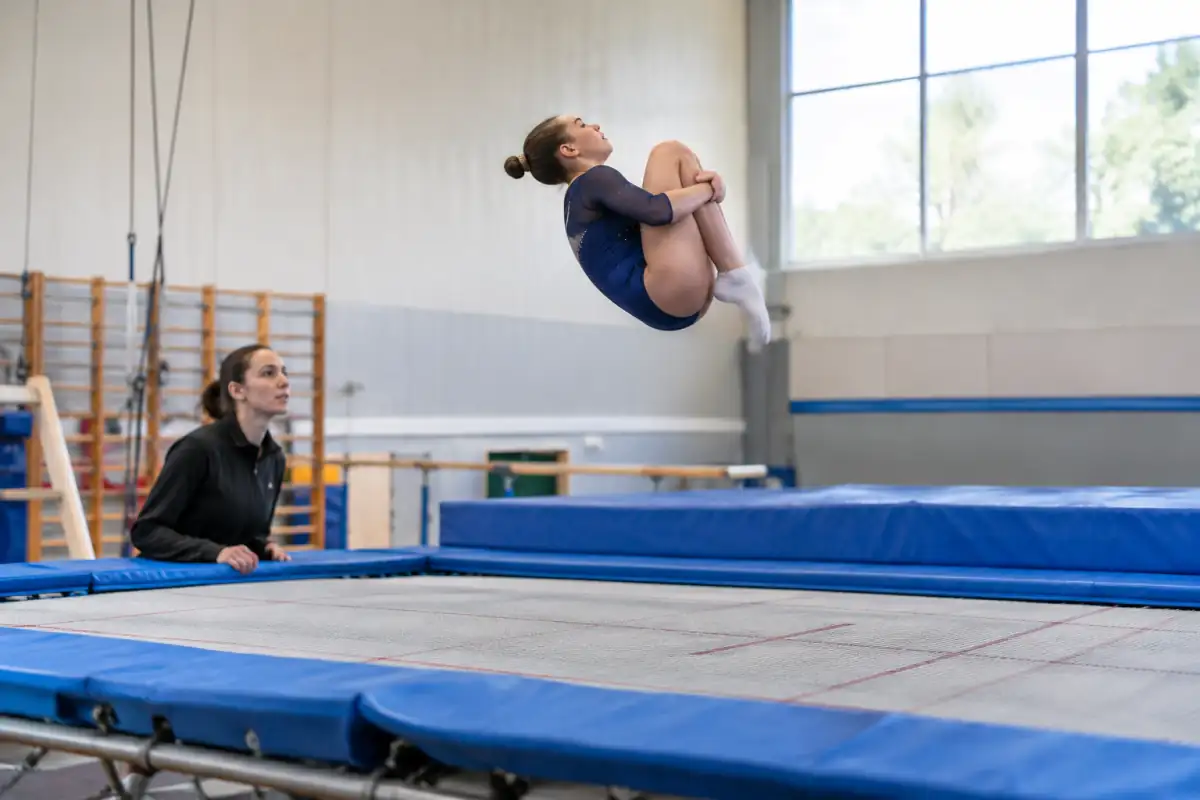 Une gymnaste réalise une rotation groupée sur un trampoline de compétition, sous le regard d’un entraîneur dans une salle équipée de tapis.