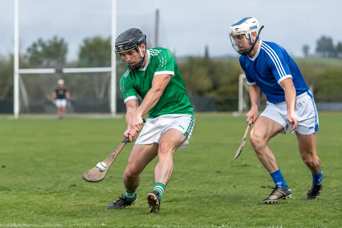 Deux joueurs casqués disputent un sliotar au hurley sur un terrain de hurling, près d’un but en H.